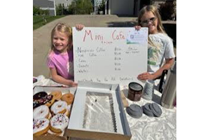 Two girls holding up a mini café sign with coffee and donuts on a table in front of them.